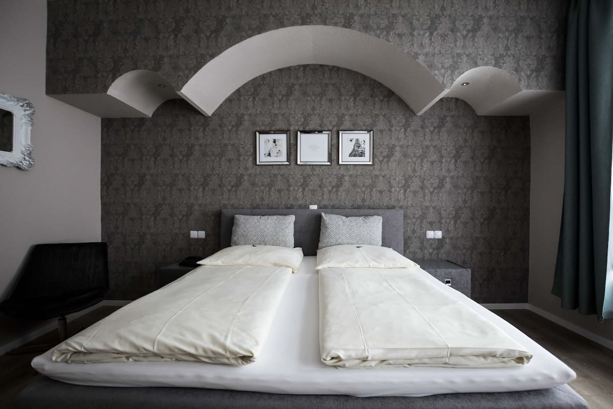 Uma cama de casal com edredom branco, duas almofadas, cabeceira e mesa de cabeceira. Há três quadros na parede acima da cama.
