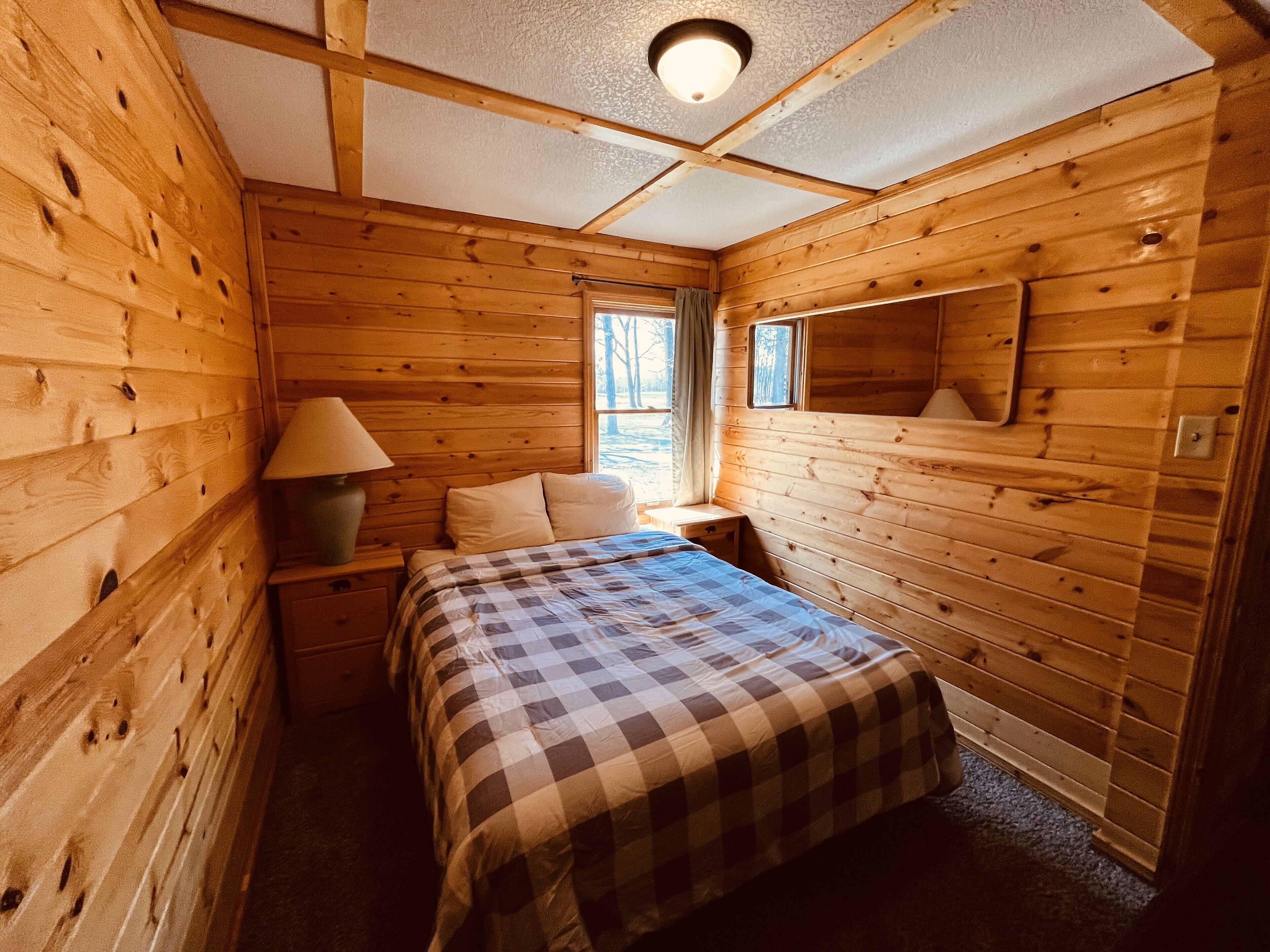 A cozy cabin room with a bed, a nightstand, a lamp, and a window with curtains.