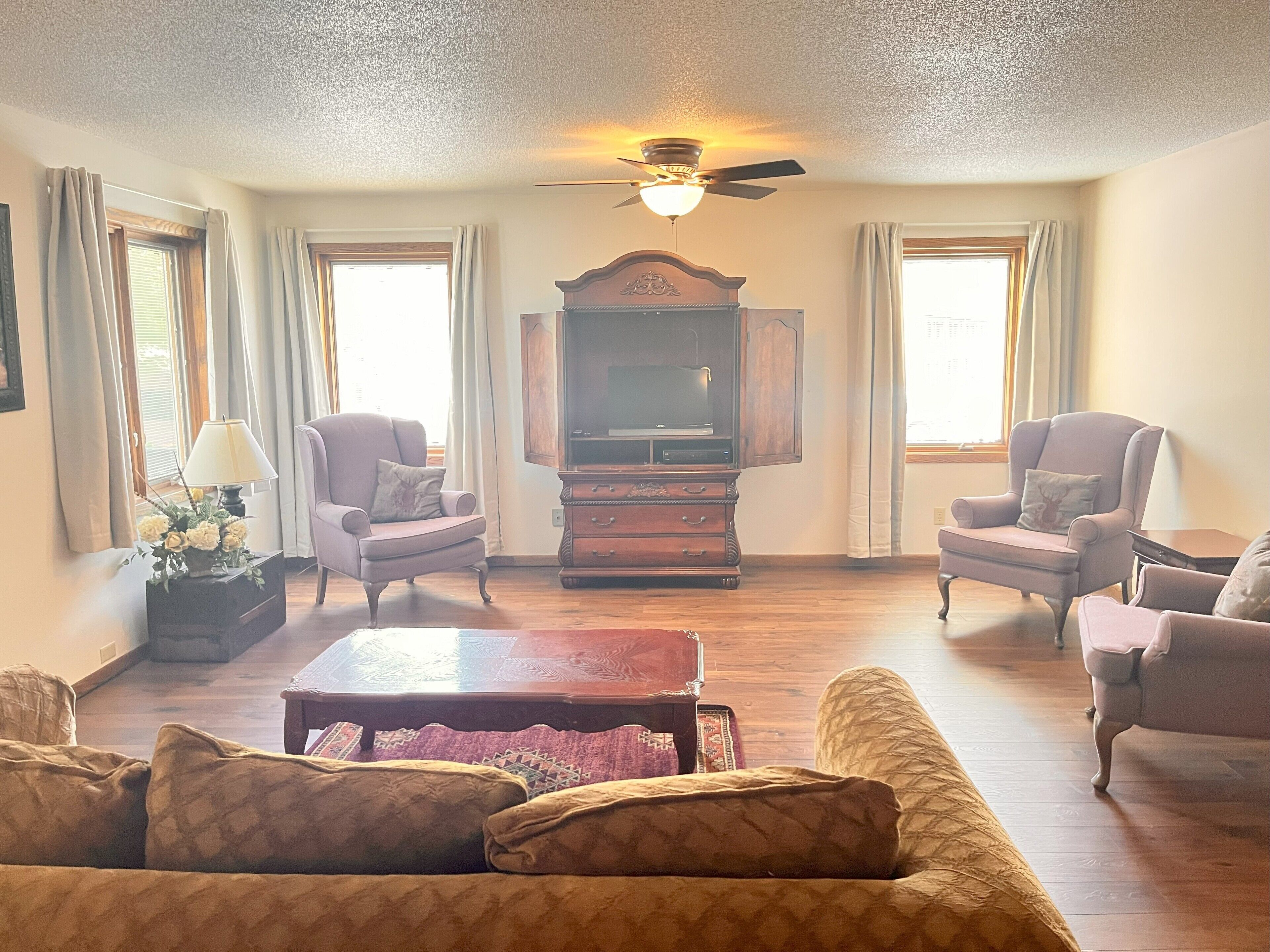 A living room with a wooden coffee table, two armchairs, a sofa, a television on a wooden cabinet, and a ceiling fan.