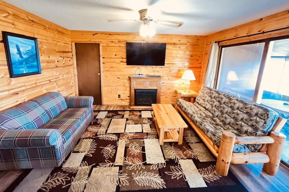 A cozy cabin interior with a fireplace, two sofas, a television, and a ceiling fan.