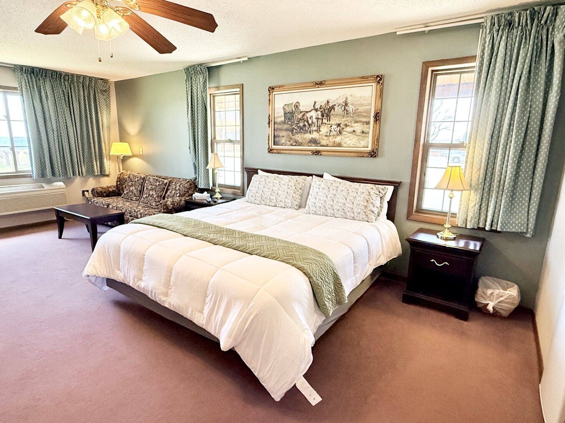 A bedroom with a large bed, a ceiling fan, a painting, and a window with curtains.