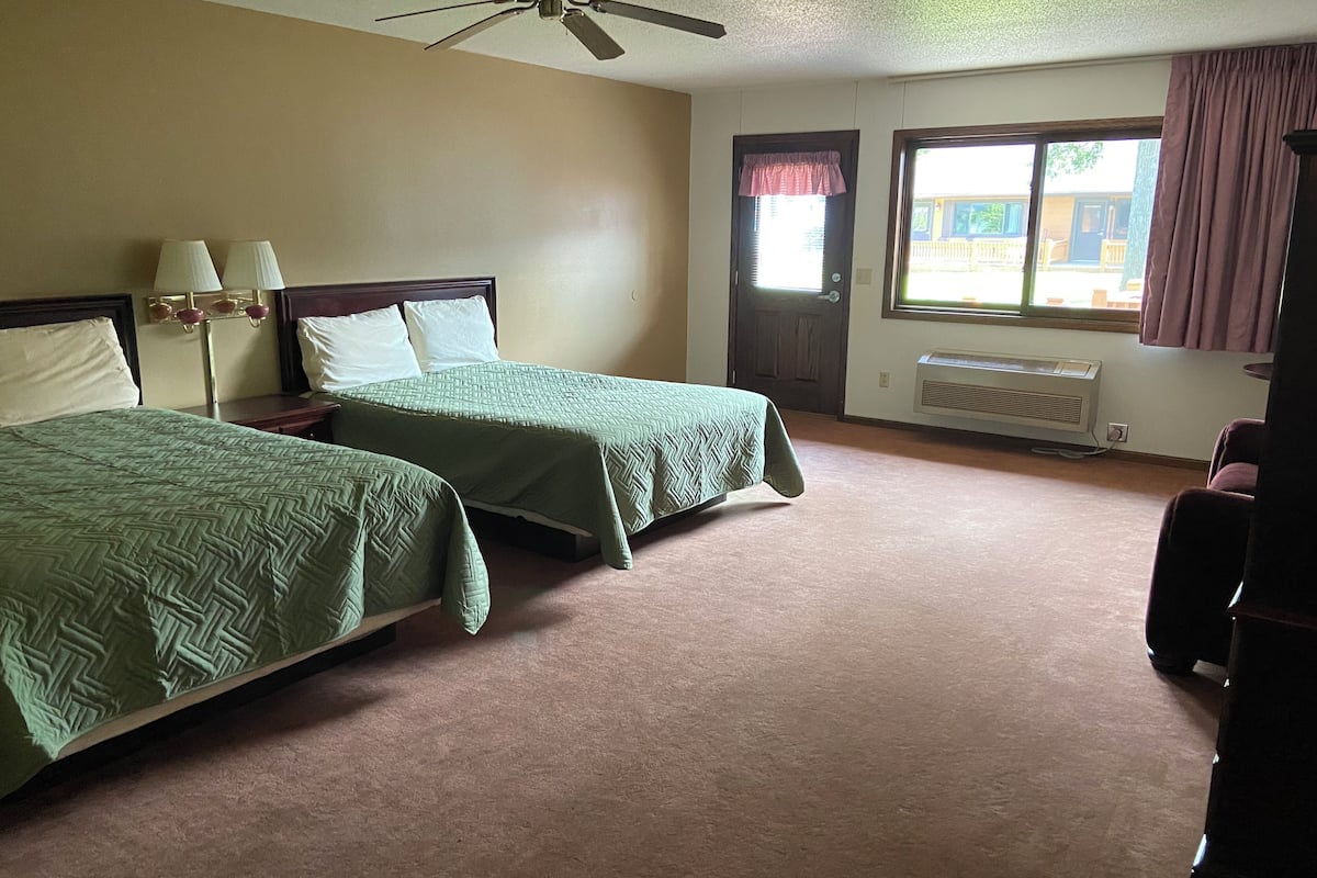 A hotel room with two beds, a ceiling fan, a window with curtains, and a radiator.