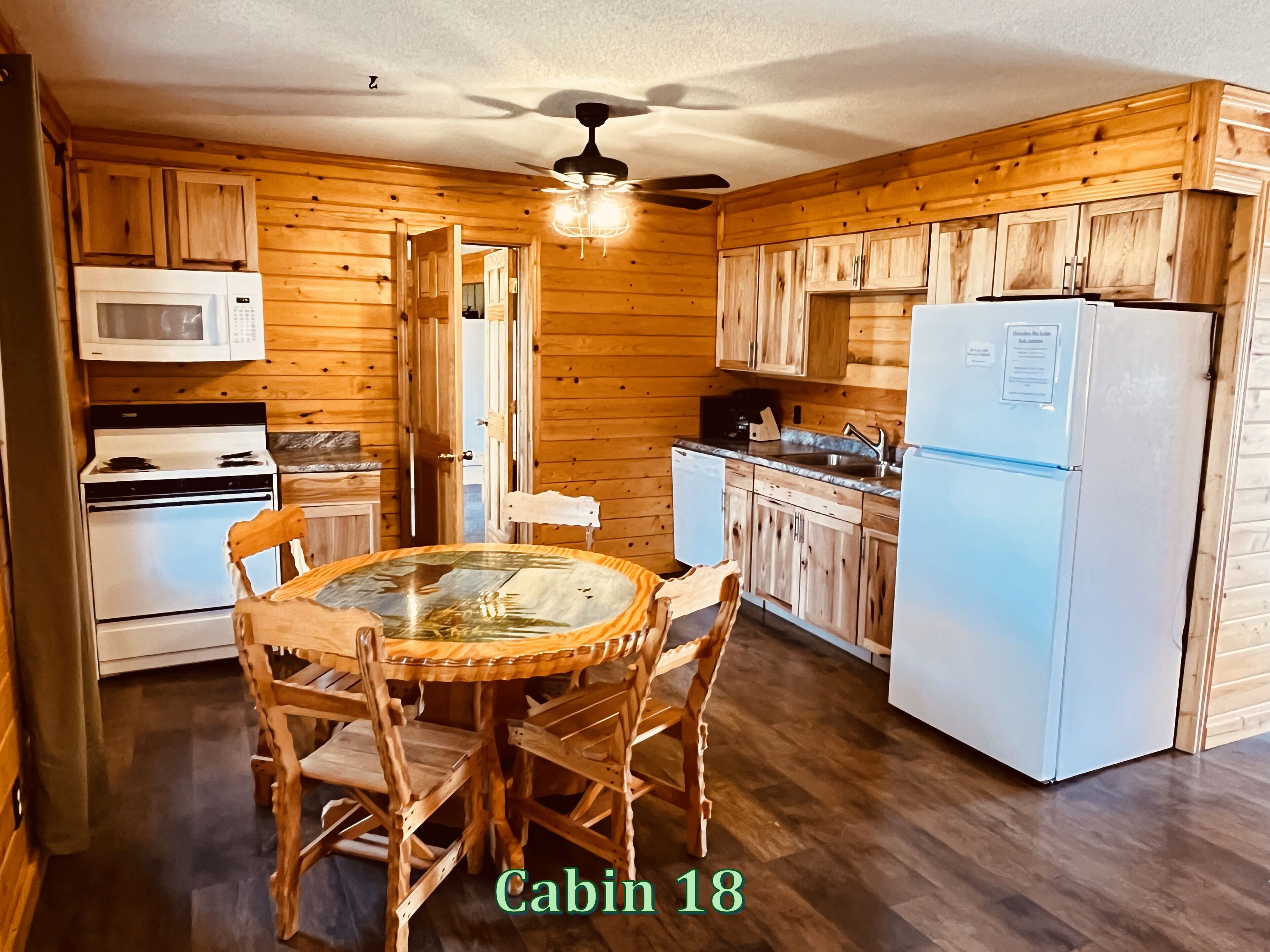 A cabin kitchen with wooden walls, a round table with chairs, a microwave, a stove, a refrigerator, and a ceiling fan.