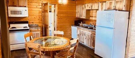 A cabin kitchen with wooden walls, a round table with chairs, a microwave, a stove, a refrigerator, and a ceiling fan.