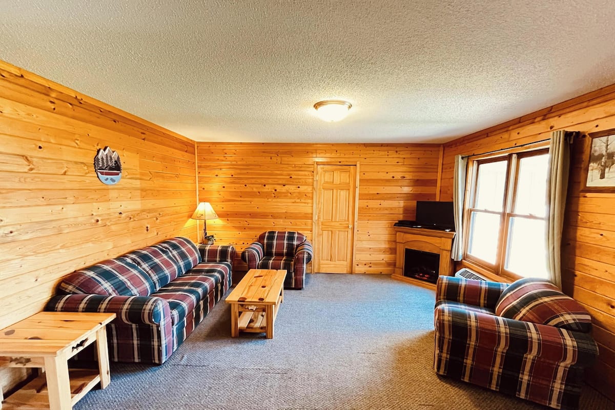 A cabin interior with a plaid sofa set, a wooden coffee table, and a television on a stand.