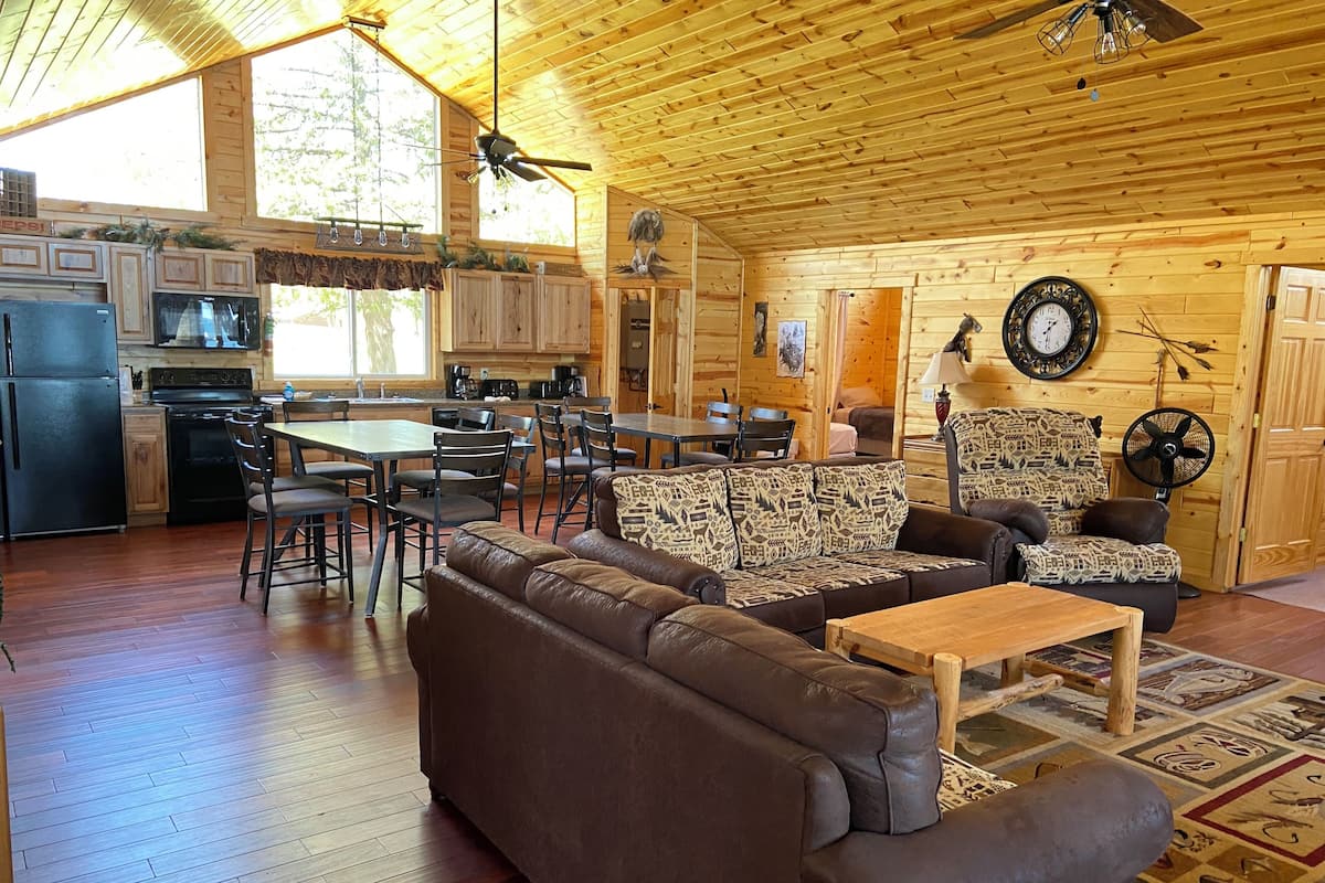 A spacious living area with a brown leather sofa, a wooden coffee table, and a dining table with chairs. The kitchen area is visible with a refrigerator, stove, and microwave. The room has wooden walls and a high ceiling with a ceiling fan.