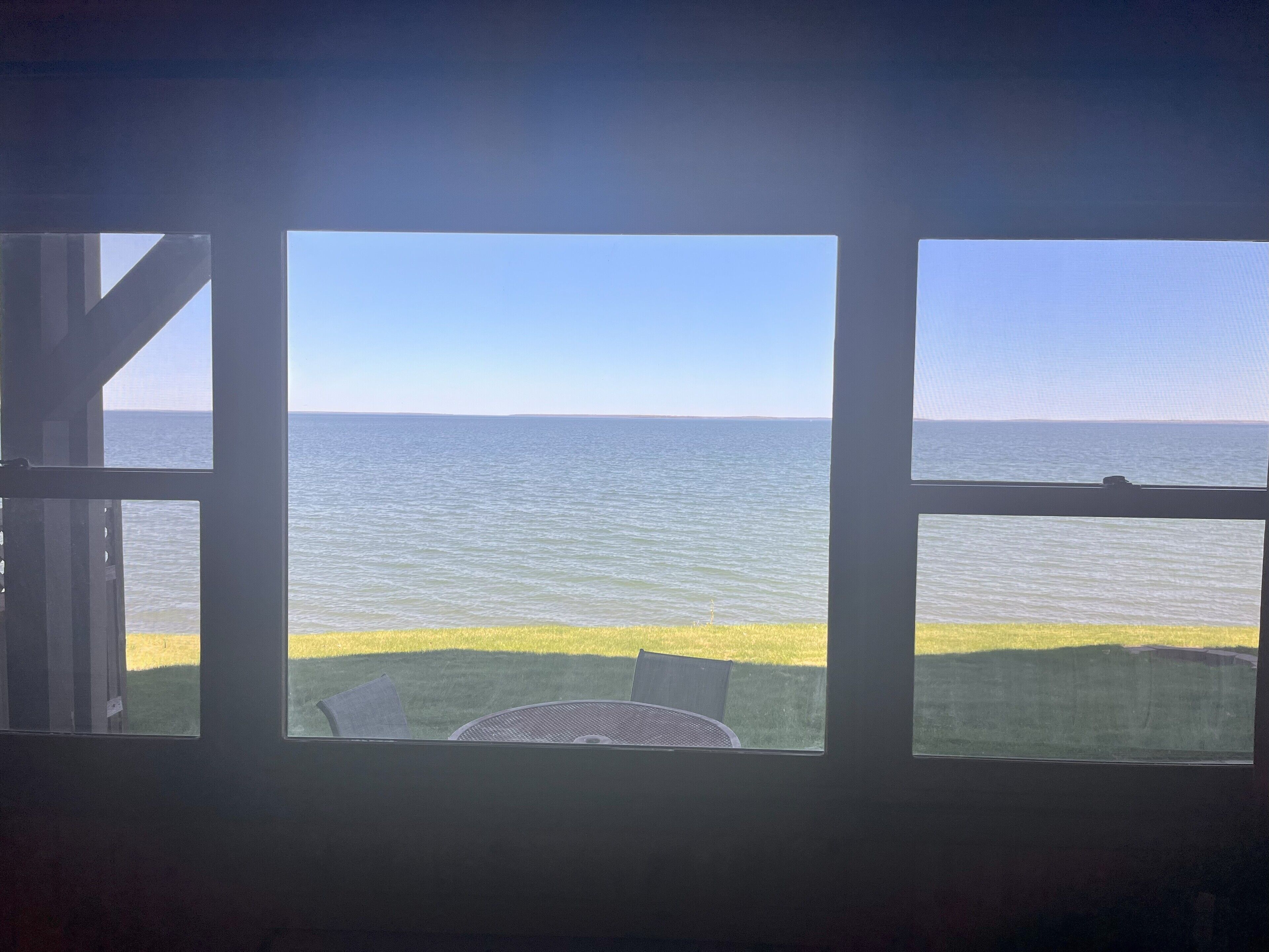 Blick durch ein Fenster auf einen Tisch und Stühle aus Metall, mit Blick auf eine Wiese und das Meer.