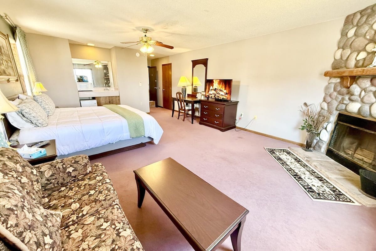 A bedroom with a bed, a wooden dresser, a desk, and a fireplace.