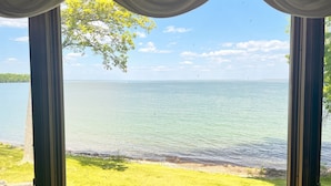 A view through a window with curtains, overlooking a calm lake, grassy area, and distant trees. - Horseshoe Bay Lodge (Walker)