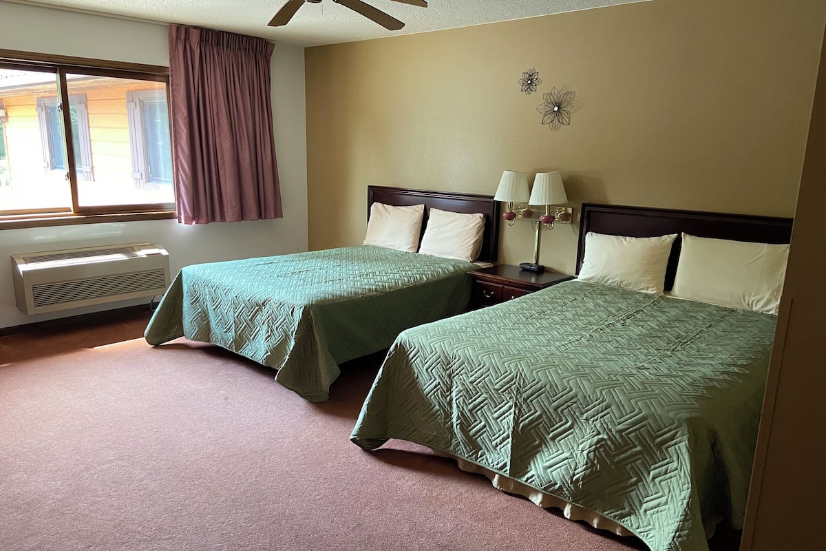 A hotel room with two beds, a window with curtains, a ceiling fan, a nightstand with a lamp, and a wall-mounted air conditioner.