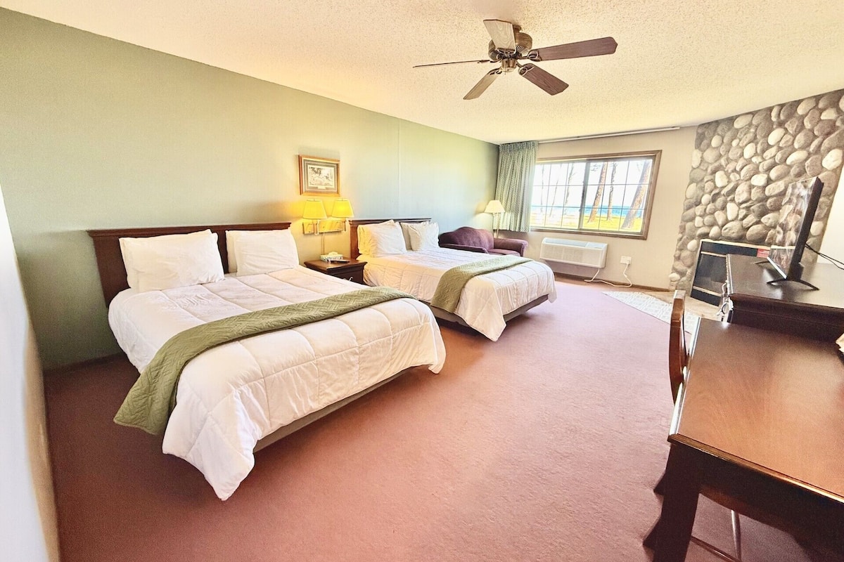 A hotel room with two beds, a desk, a ceiling fan, a window with a view, and a stone wall.