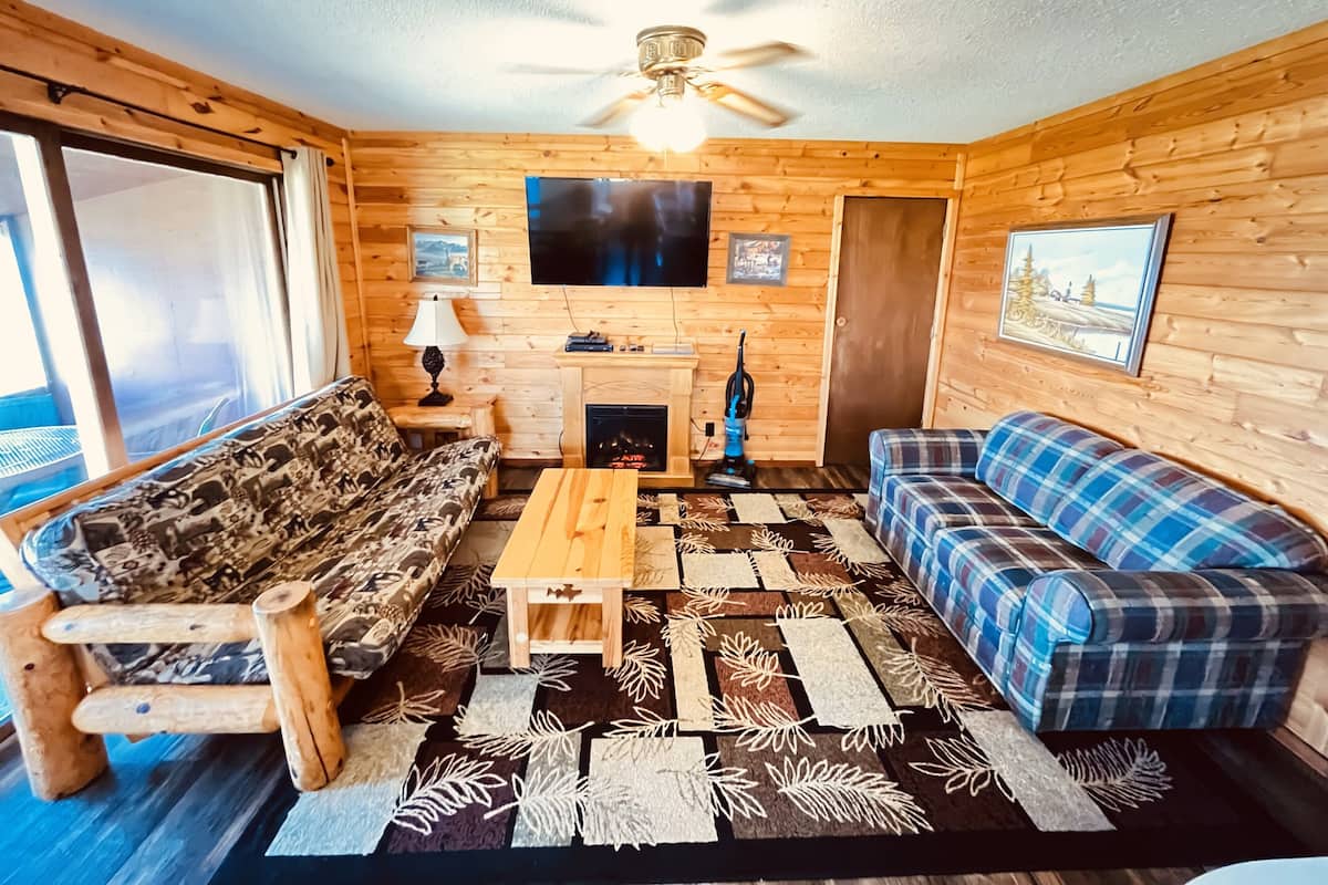 A cozy cabin-style room with wooden walls, a ceiling fan, a fireplace, a television, and two sofas.