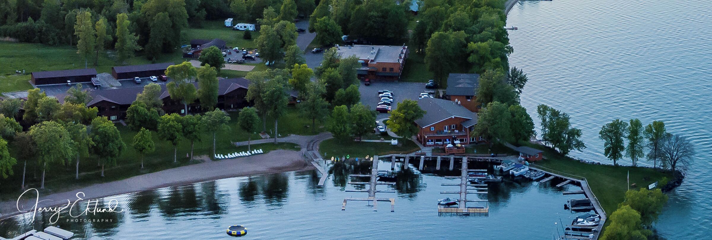 An aerial view of a lakeside property with multiple buildings, a dock with boats, and a parking area.