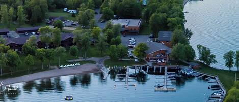 An aerial view of a lakeside property with multiple buildings, a dock with boats, and a parking area.