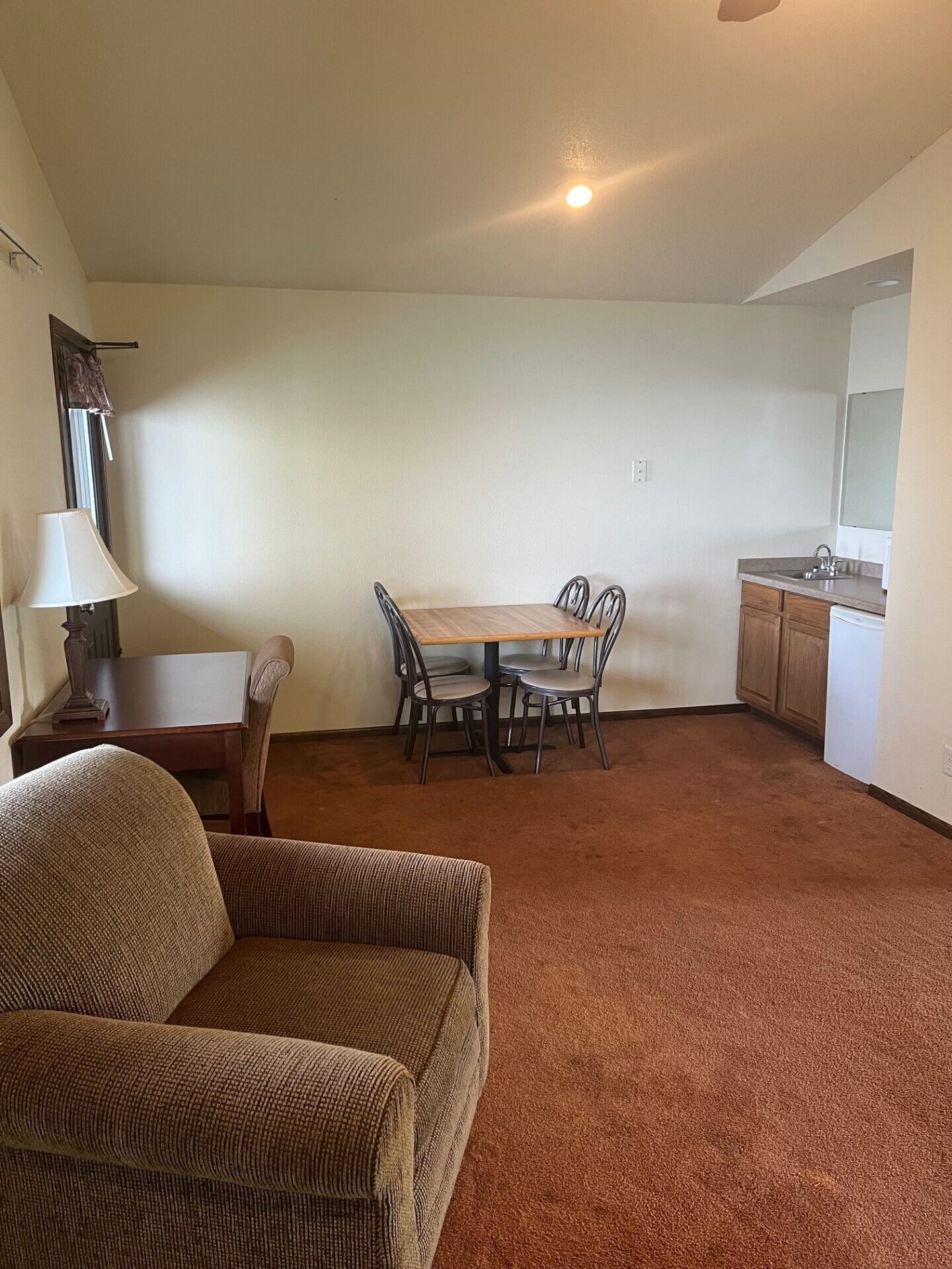 A compact living space with a dining table, four chairs, a sofa, a lamp, a small cabinet, and a kitchen area with a sink.