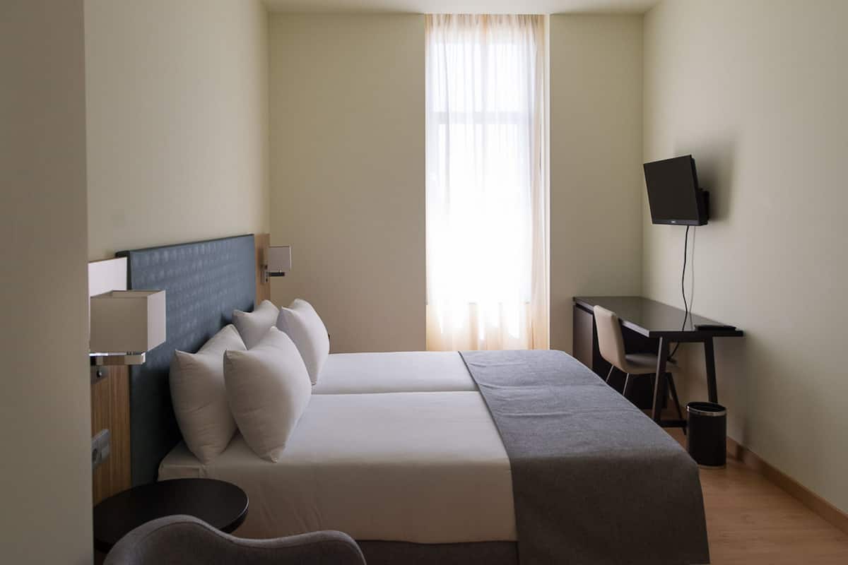 A hotel room with a bed, a desk, a chair, a TV, and a window with curtains.