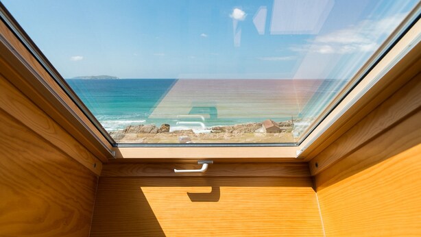 A room with a skylight, wooden walls, and a view of the ocean and rocky shore. - Pousada A Lanzada (O Grove)