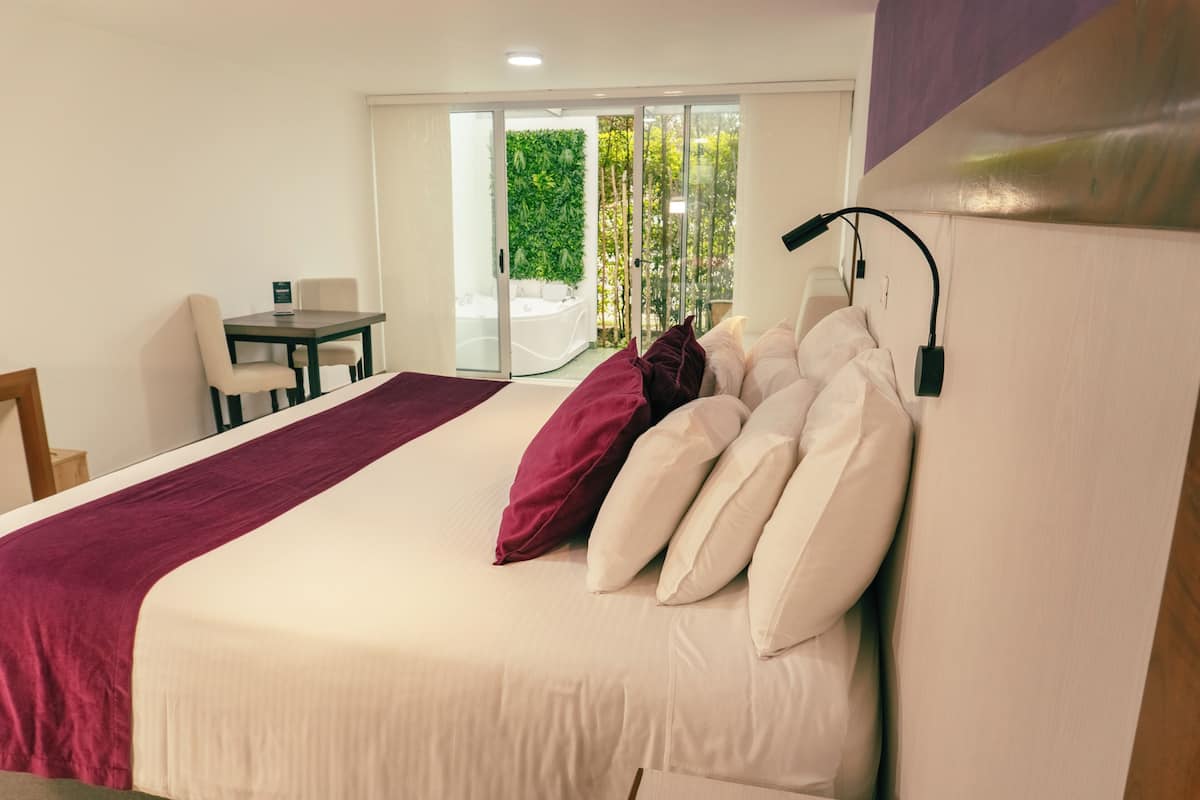 A hotel room with a bed, a desk, and a chair. There is a sliding glass door leading to a balcony with a view of greenery.