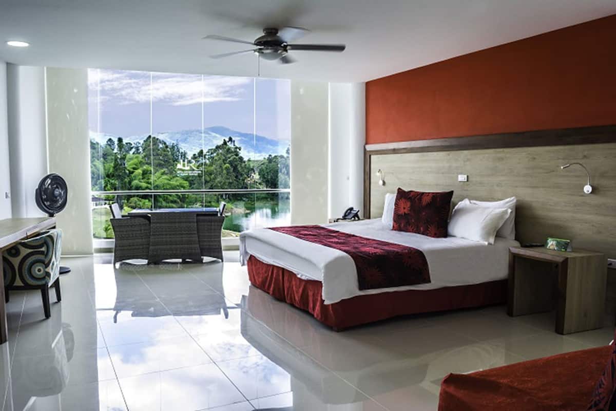 A modern hotel room with a large bed, a desk, a chair, and a balcony with a view of greenery and mountains.