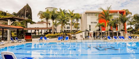 A swimming pool with blue and white tiled patterns, surrounded by lounge chairs and palm trees, with a building featuring a distinctive rock formation in the background.