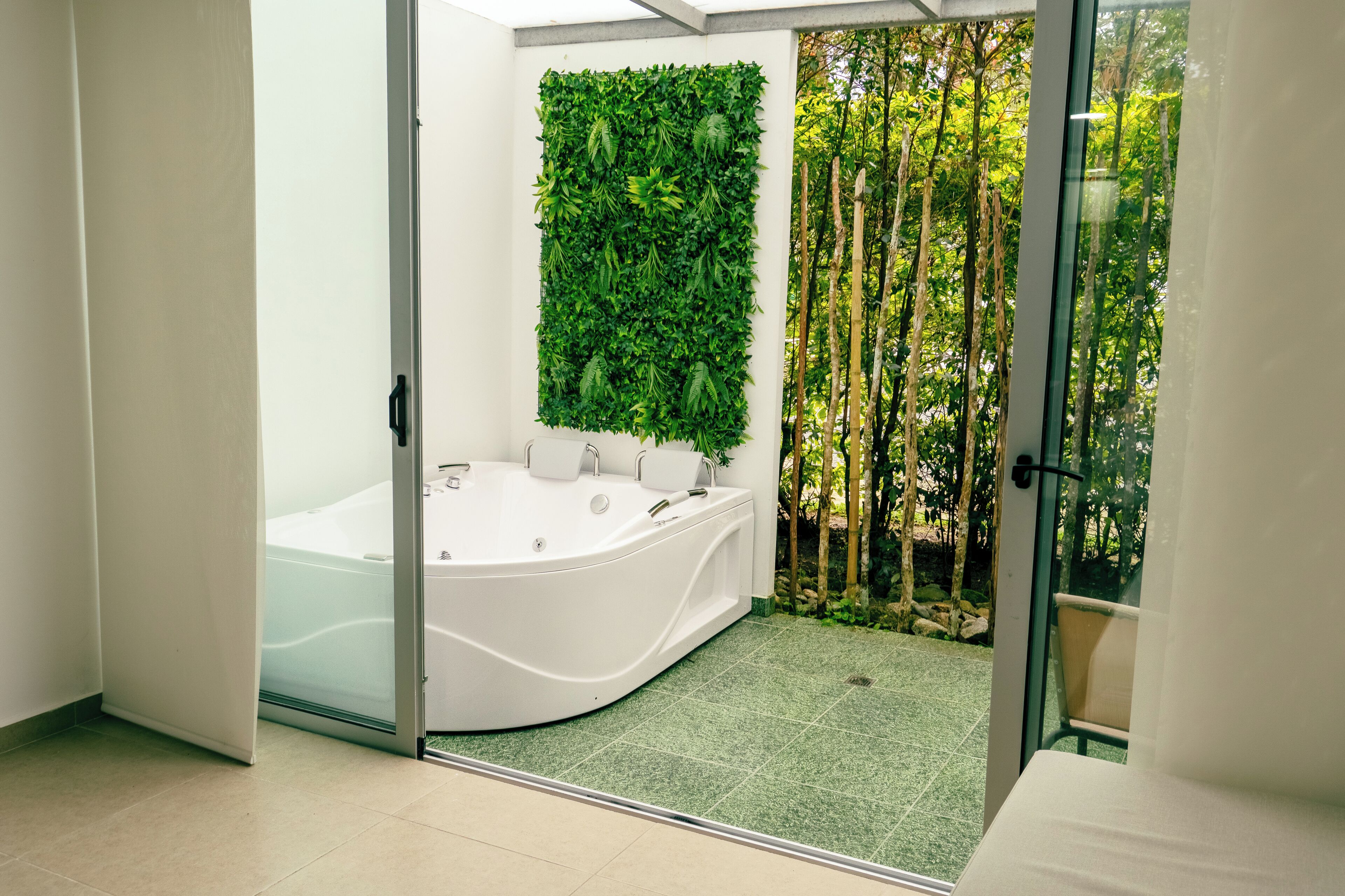 A modern bathroom with a large freestanding bathtub, a green vertical garden wall, and a view of a forest through a glass door.