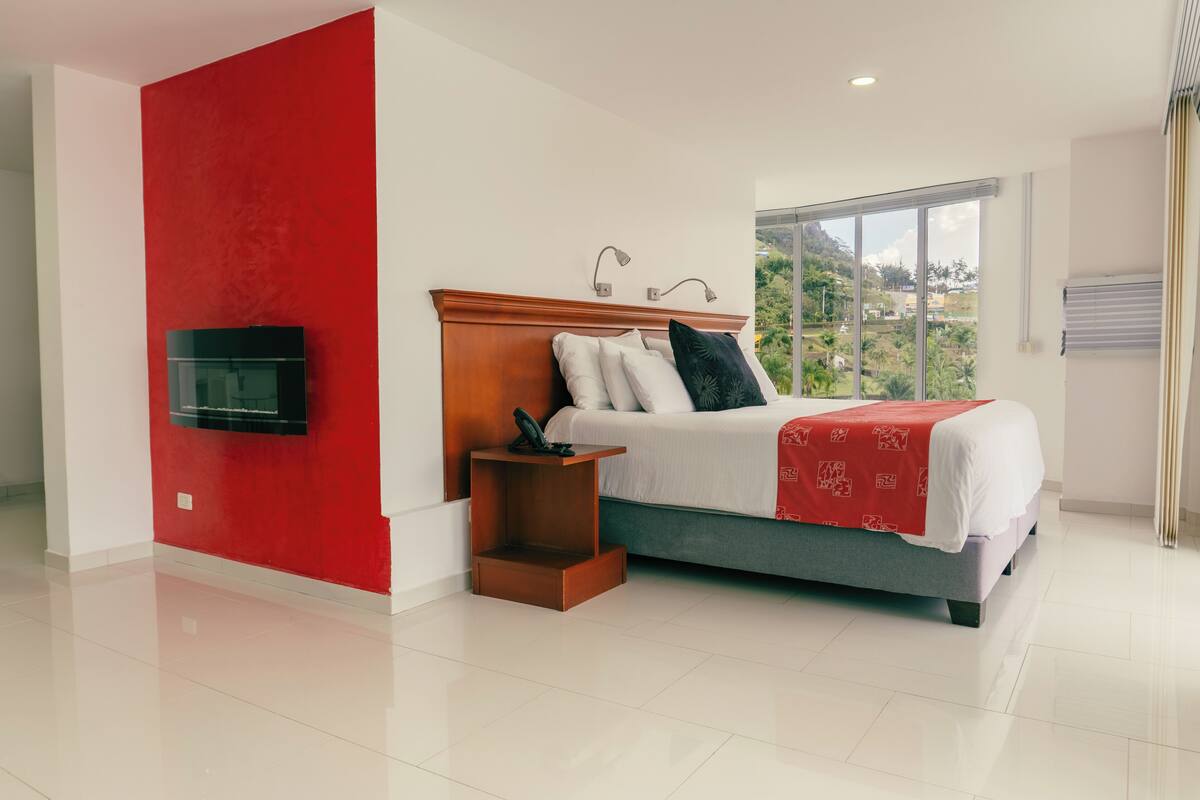 A modern bedroom with a bed, a TV mounted on the wall, and a window with a view of greenery.