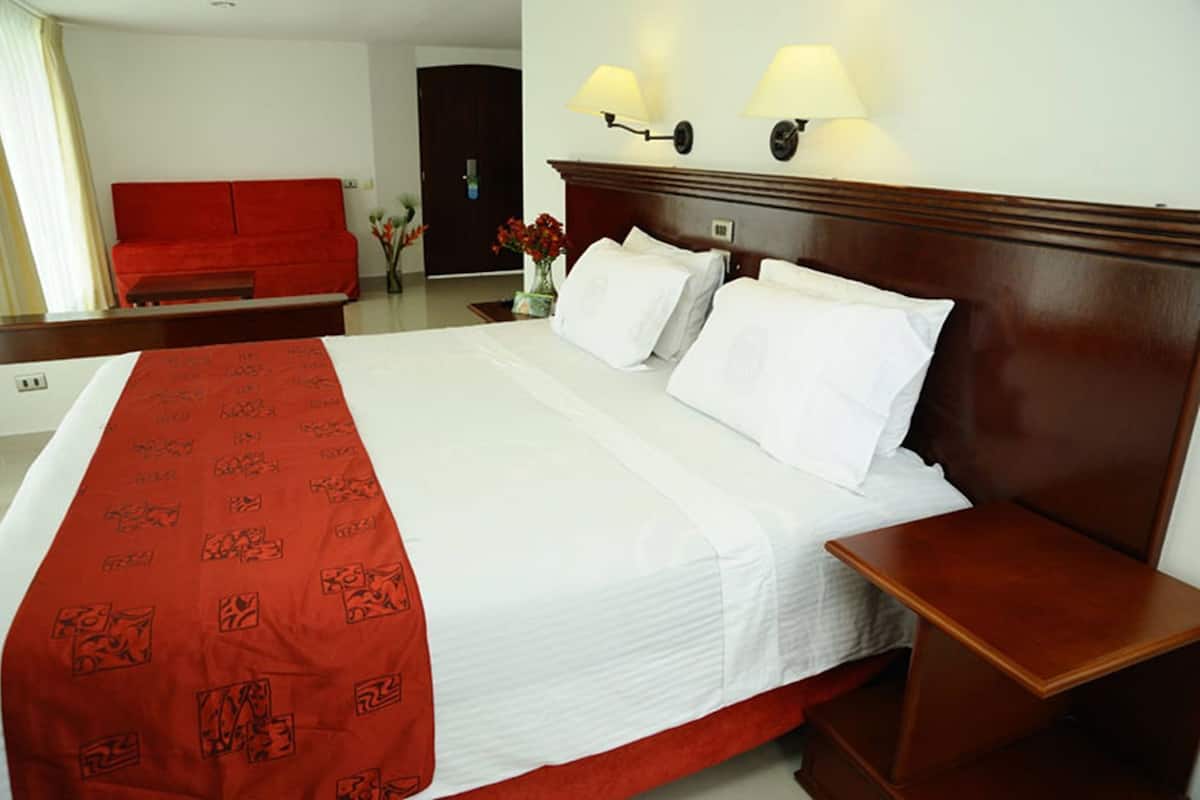 A hotel room with a large bed, two bedside tables, a red sofa, and a door in the background.
