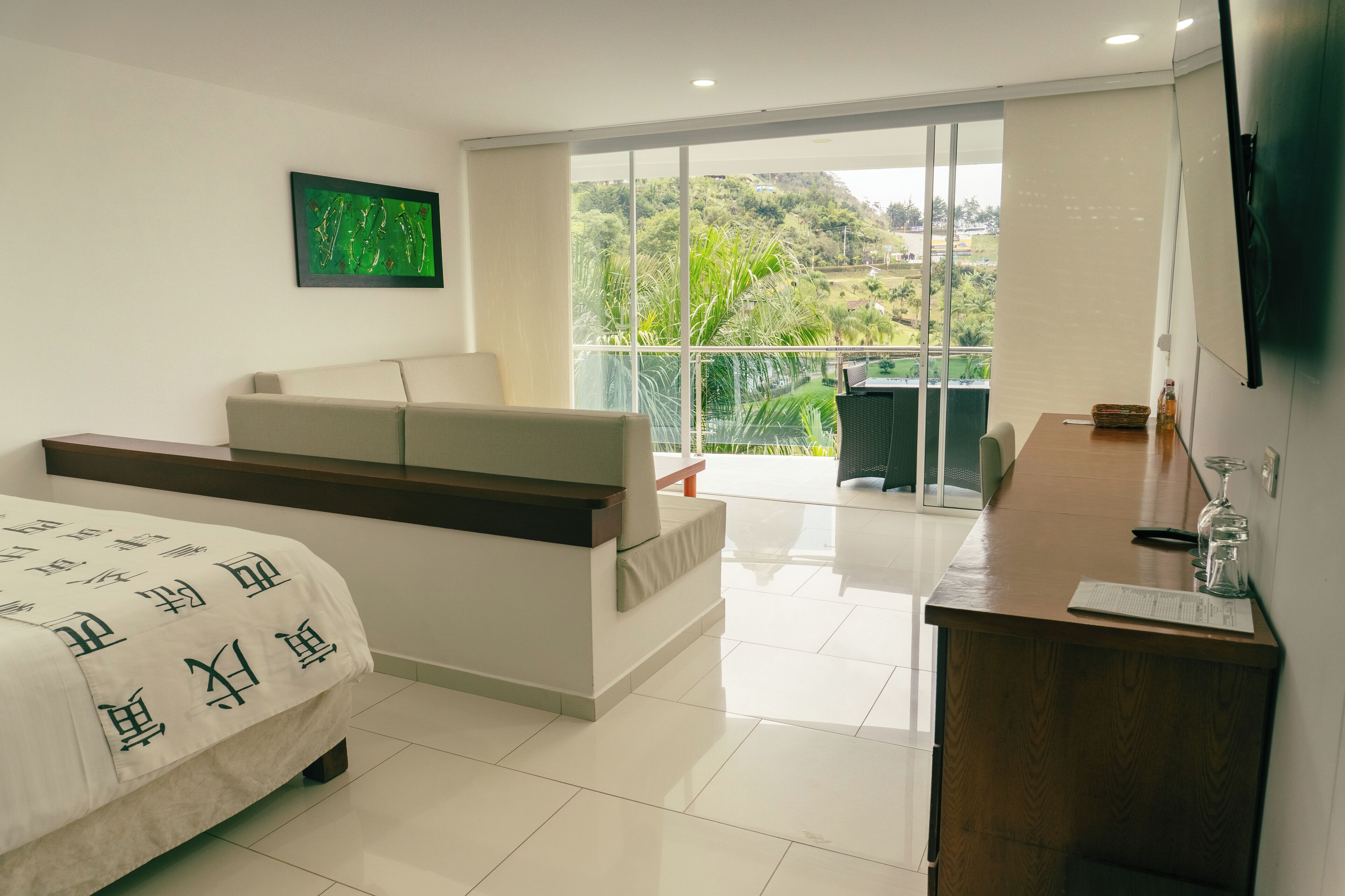 Un dormitorio moderno con cama, sofá, balcón con vistas a la vegetación y un escritorio de madera con un vaso.