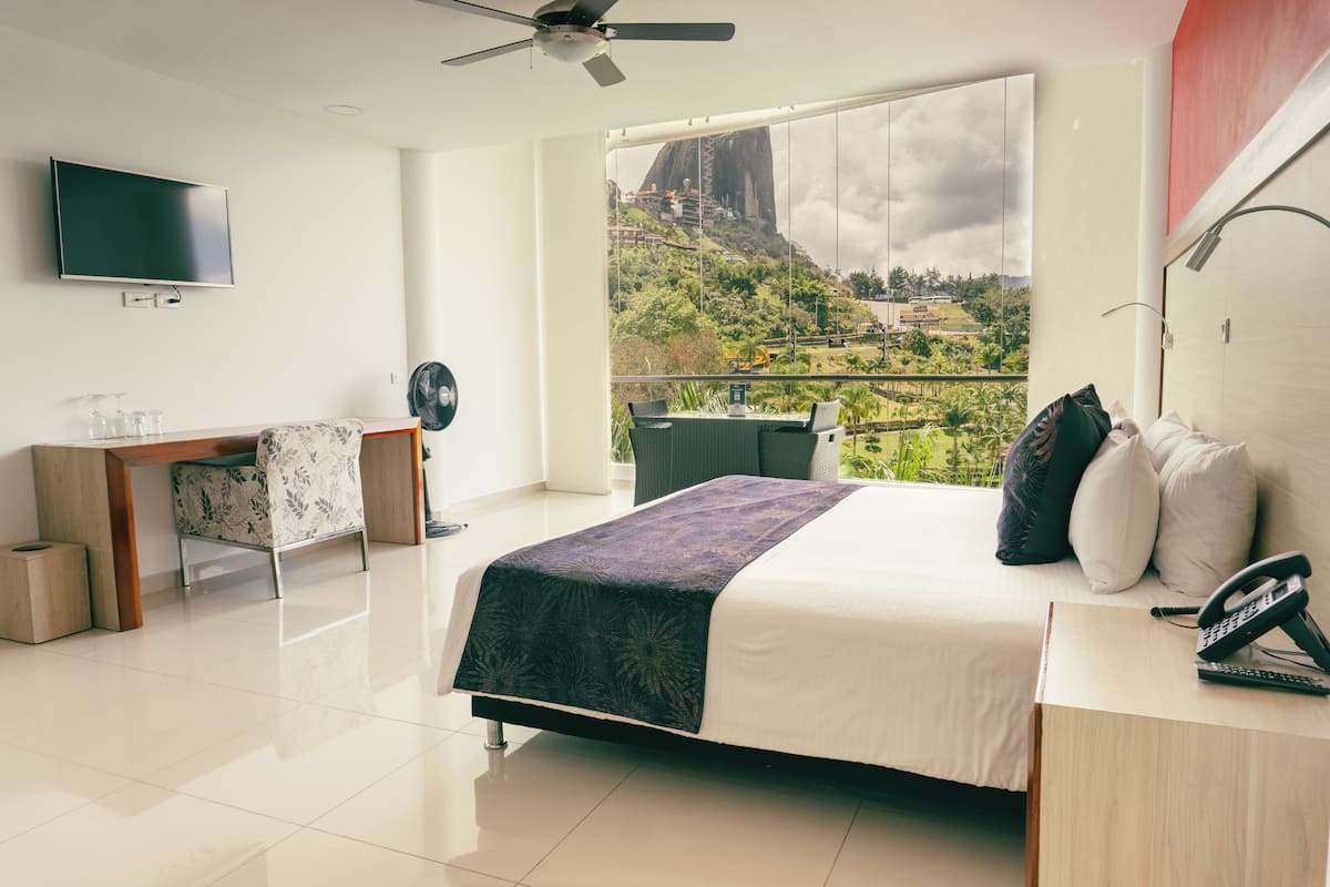 A hotel room with a bed, a desk, a chair, a TV, a balcony with a view of a mountain and clouds.