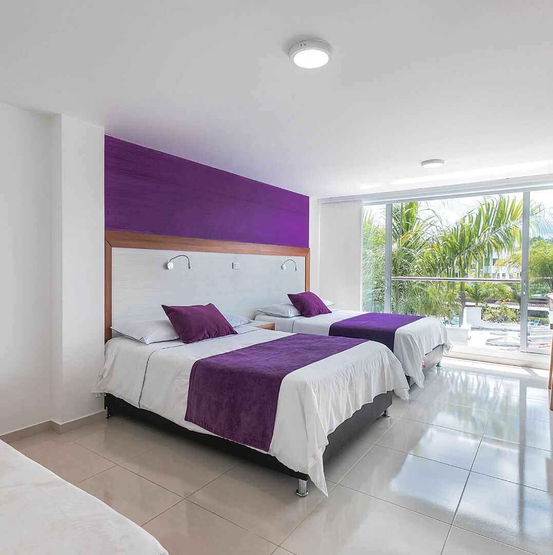A modern hotel room with two beds, a large window with a view of palm trees, and a bright ceiling light.