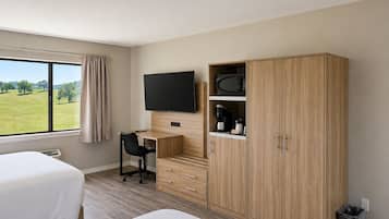 A hotel room with two beds, a desk with a chair, a TV, a wardrobe, and a view of a green field.
