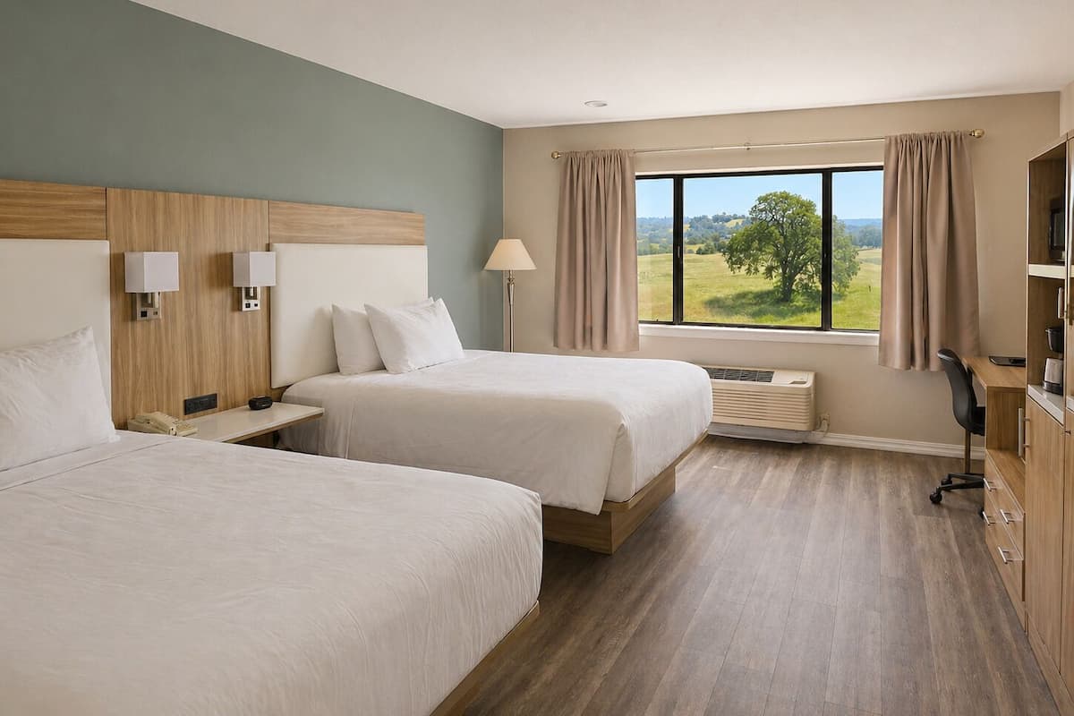 A hotel room with two beds, a desk, a chair, a lamp, and a view of the outdoors.