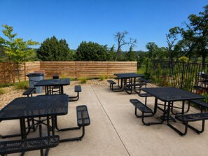 Un espace extérieur aménagé en terrasse, avec des tables de pique-nique et des bancs, entouré d’une clôture en bois et de verdure.