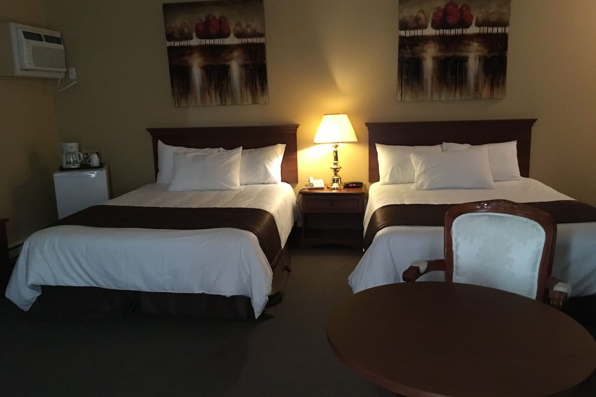 A hotel room with two beds, a bedside table with a lamp, a coffee maker, and two paintings on the wall.