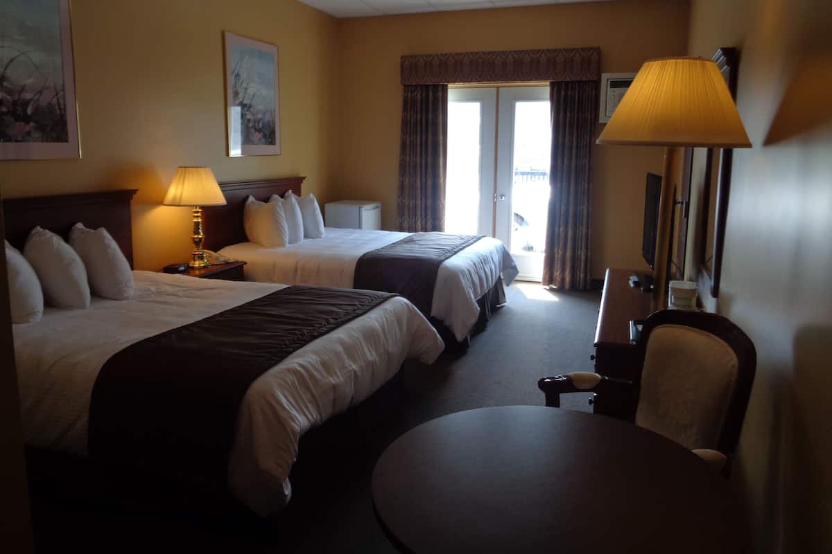 A hotel room with two beds, a desk, a chair, a lamp, and a large window with curtains.