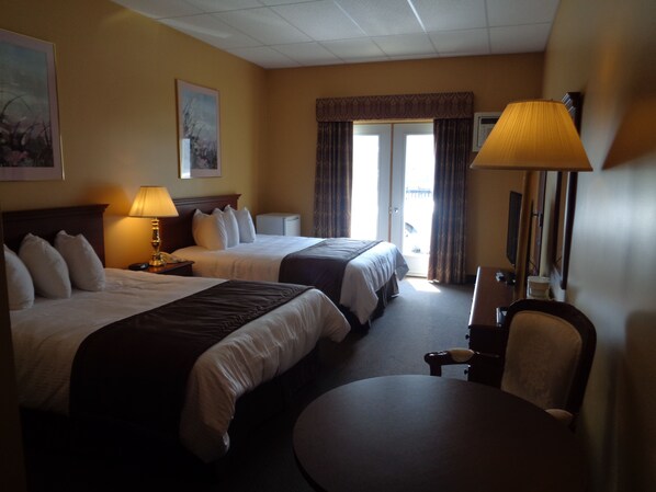 A hotel room with two beds, a desk, a chair, a lamp, and a large window with curtains. - Motel Cofotel (Saint-Simeon)