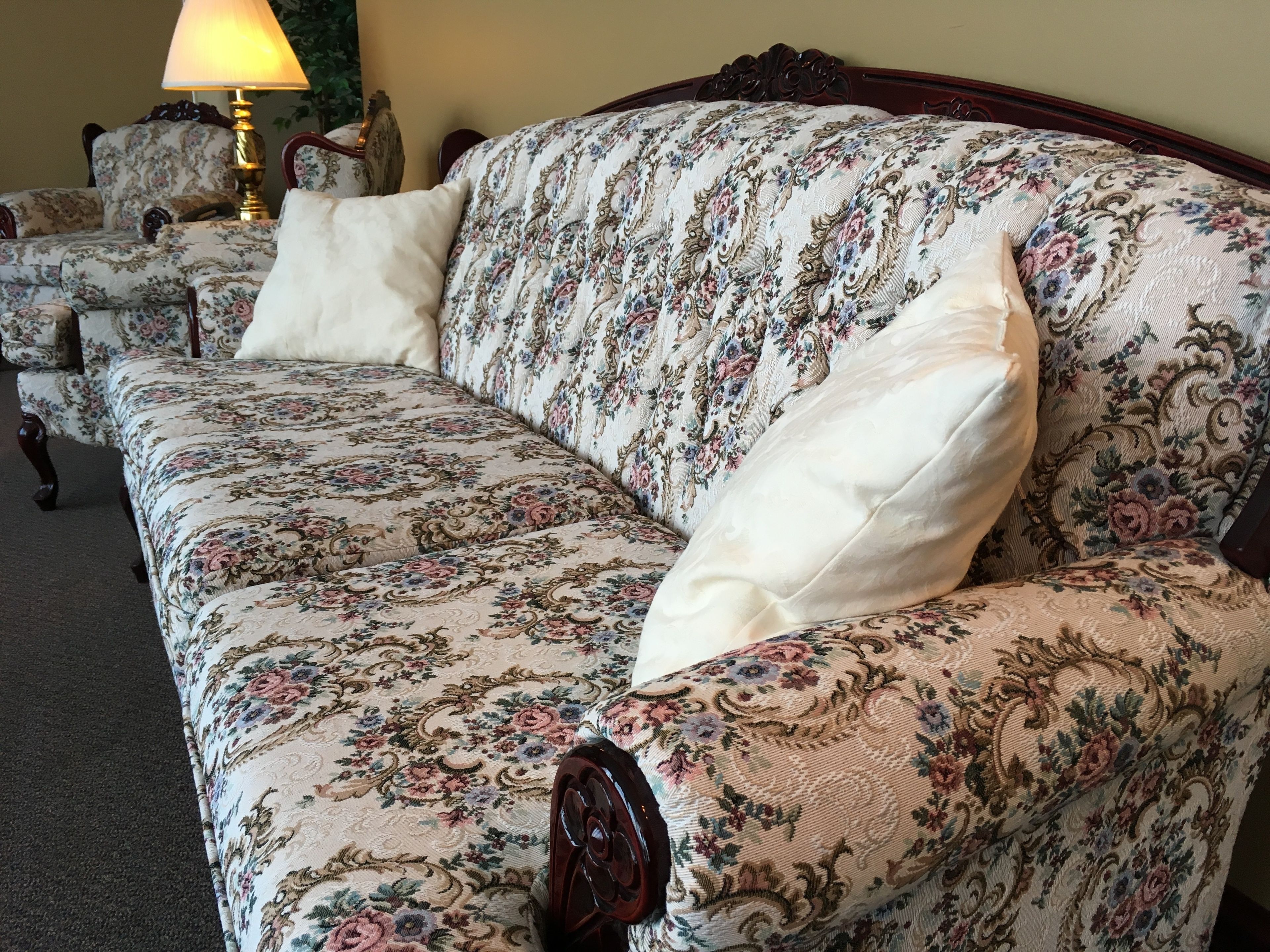 Un sofá estampado con motivos florales, con estructura de madera, acompañado por sillones a juego y una lámpara de mesa.