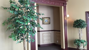 A hallway with a large artificial plant, a framed picture on the wall, and a potted plant. - Motel Cofotel (Saint-Simeon)