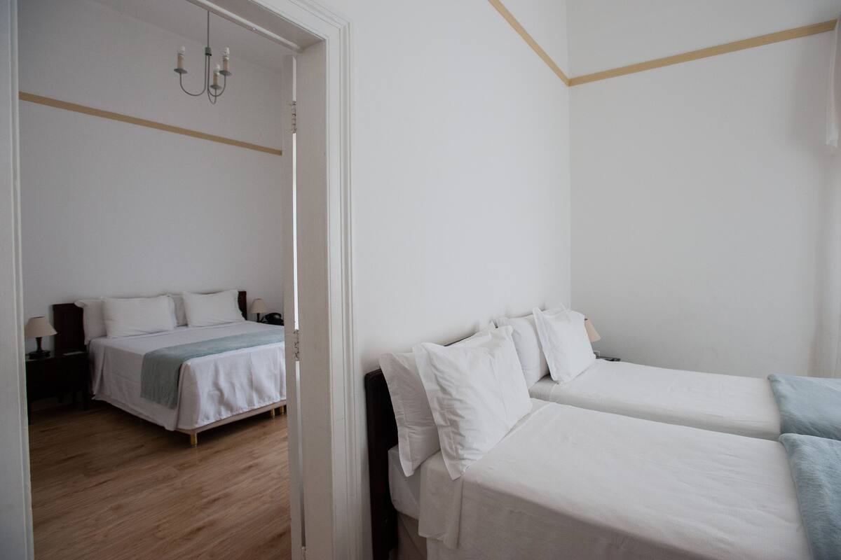 Ein Hotelzimmer mit zwei Einzelbetten, Holzboden, weißer Wand und einer Tür zu einem anderen Raum.