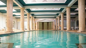 Una gran piscina interior con columnas, una zona cubierta y vistas al exterior.