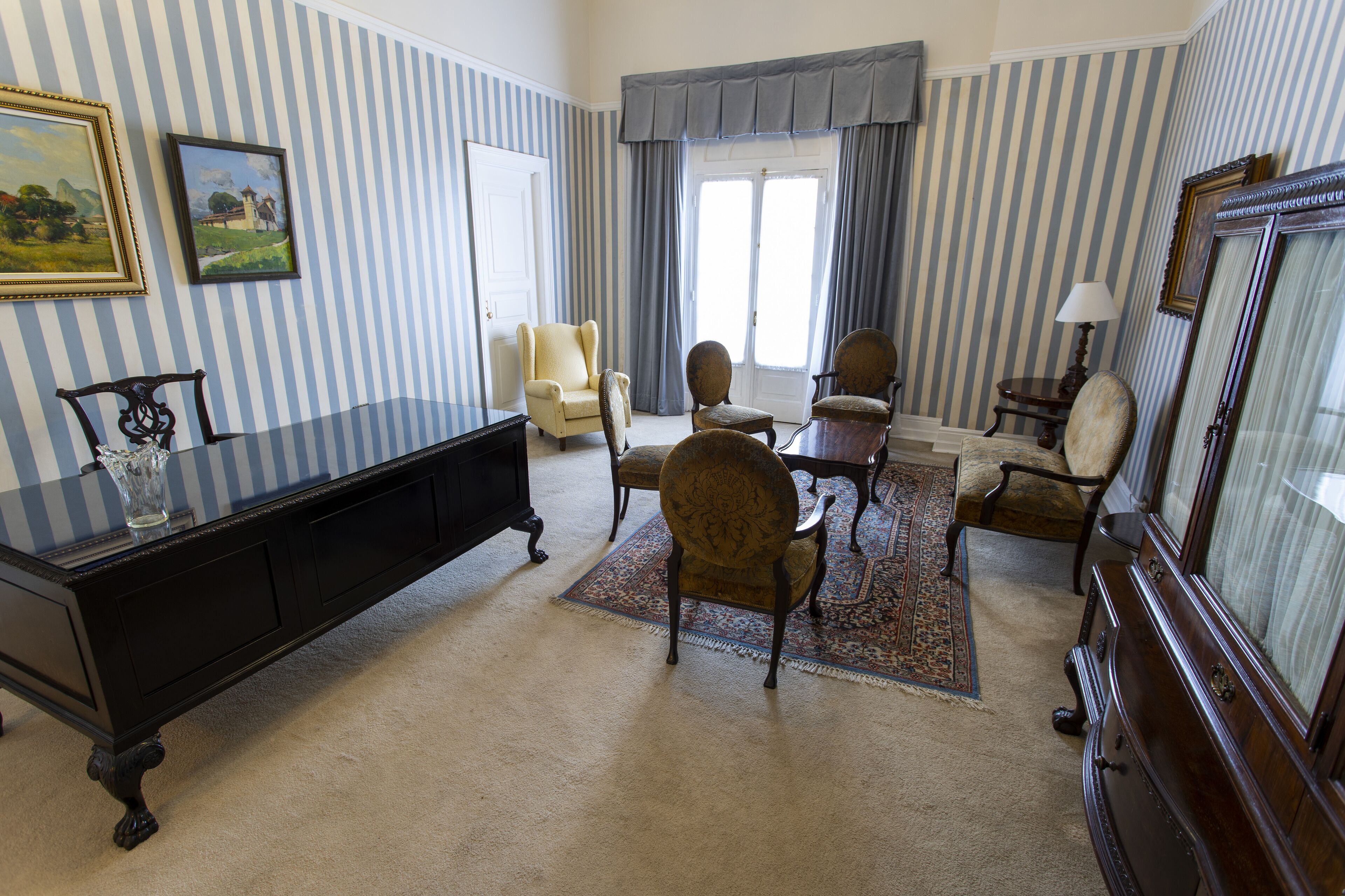 A room with striped wallpaper, a large window with curtains, a wooden cabinet, and several upholstered chairs.