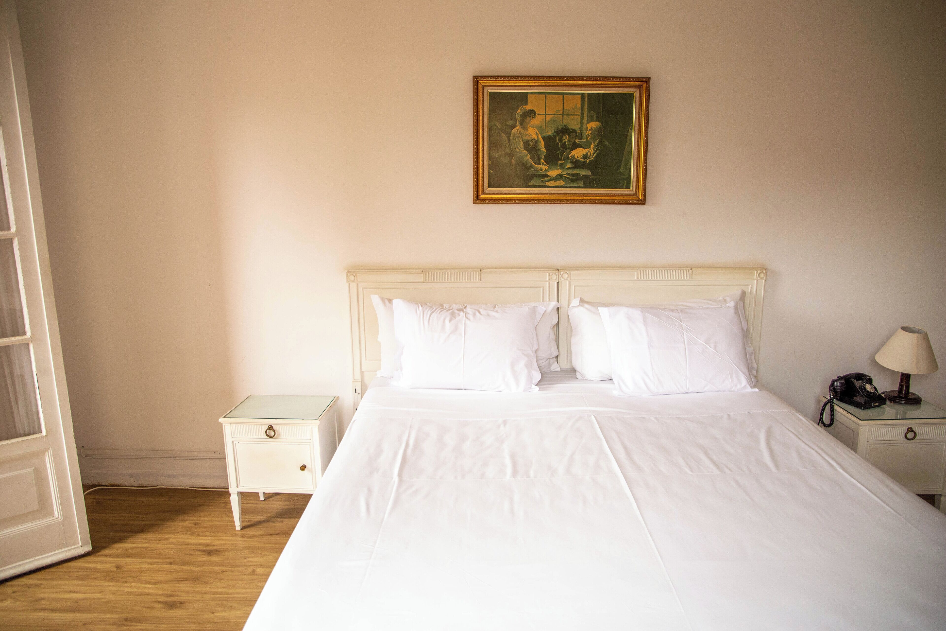 Una cama bien tendida con sábanas blancas, una mesita de noche con una lámpara y un cuadro en la pared.