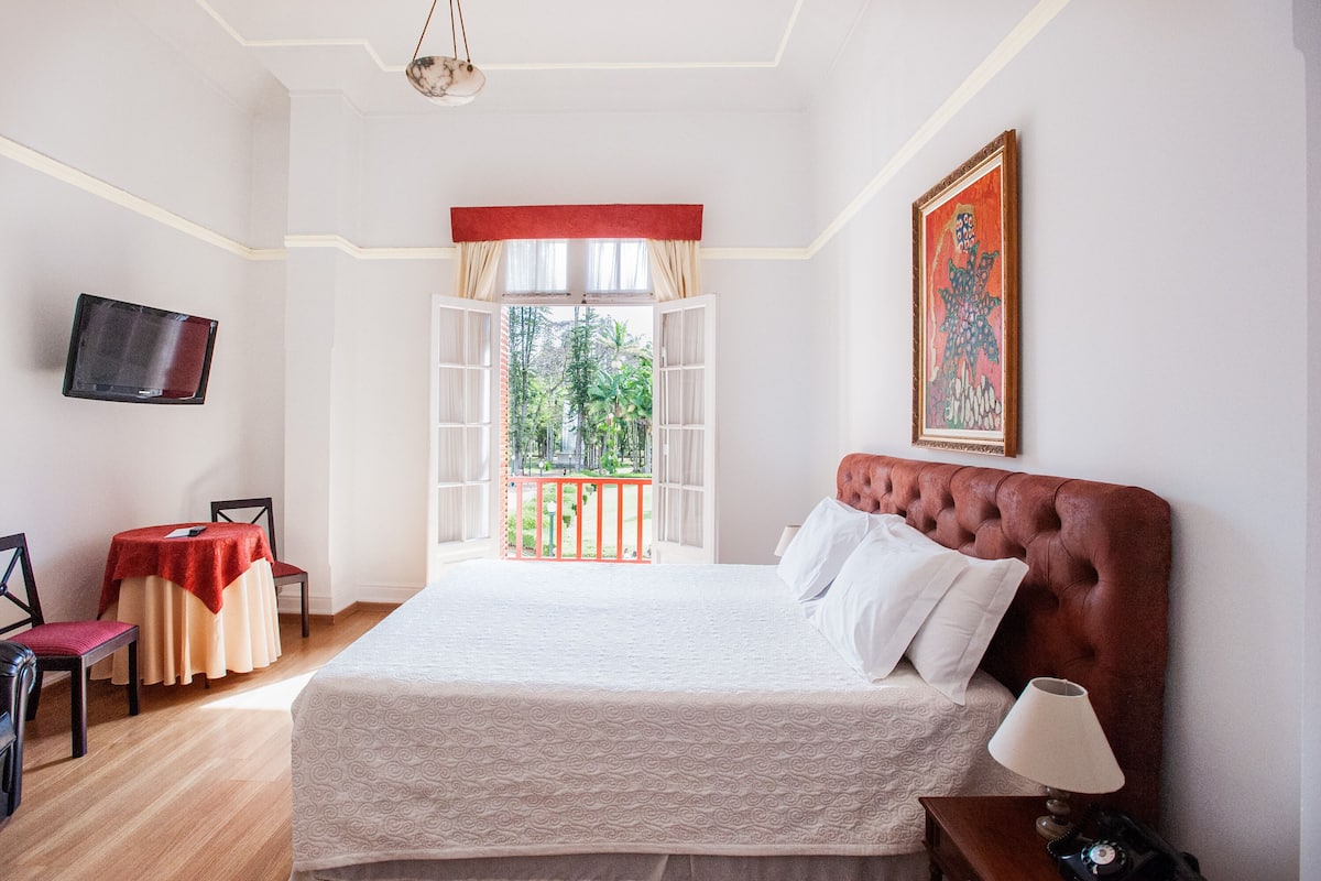 Ein Hotelzimmer mit einem großen Bett, einem an der Wand befestigten Fernseher, einem Tisch mit Stühlen und einem Balkon mit Blick auf Grünflächen.