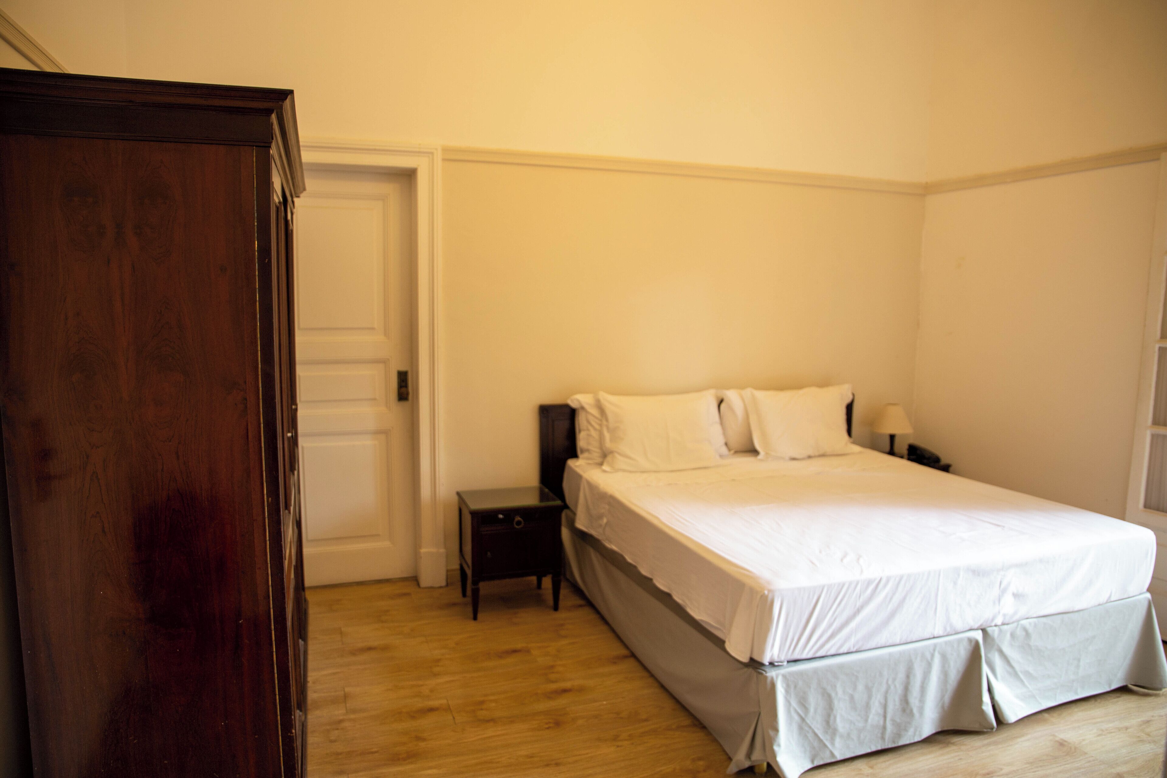 A bedroom with a wooden wardrobe, a bed with white bedding, a nightstand, and a lamp.