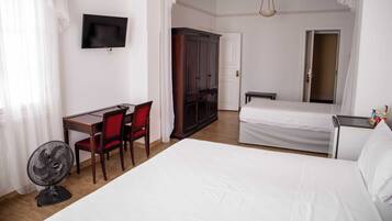 A hotel room with a bed, a desk with a chair, a wardrobe, a television, and a fan.