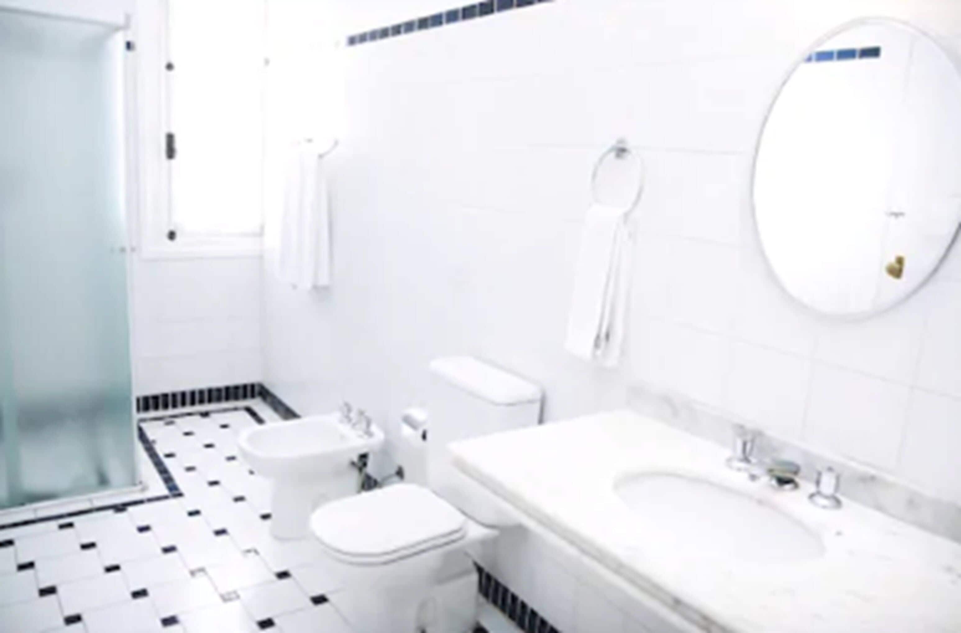 Un baño con lavamanos, inodoro y bidé blancos, suelo de baldosas y un espejo.