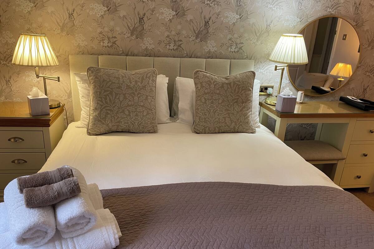 Uma cama bem arrumada com duas almofadas decorativas, uma mesa de cabeceira com abajur, um espelho e uma pilha de toalhas.