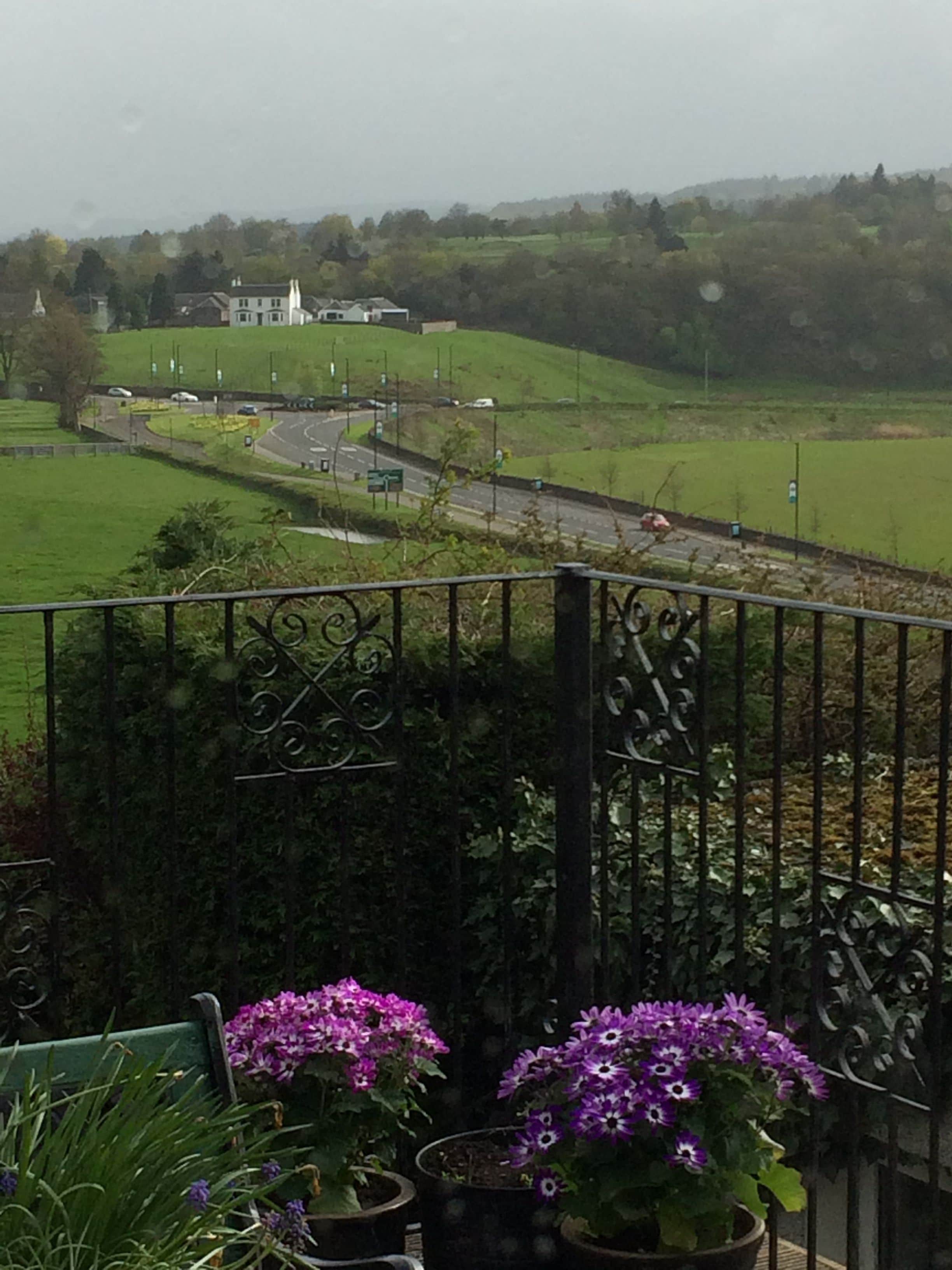 A balcony with a view of a green landscape, a road, and a building in the distance.