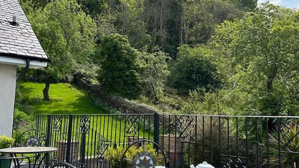 Une terrasse avec une table dressée pour le thé, entourée d’une clôture en métal, d’où l’on peut admirer un jardin luxuriant et une colline.