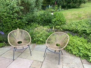 Two wicker chairs with metal frames on a stone patio surrounded by lush greenery.